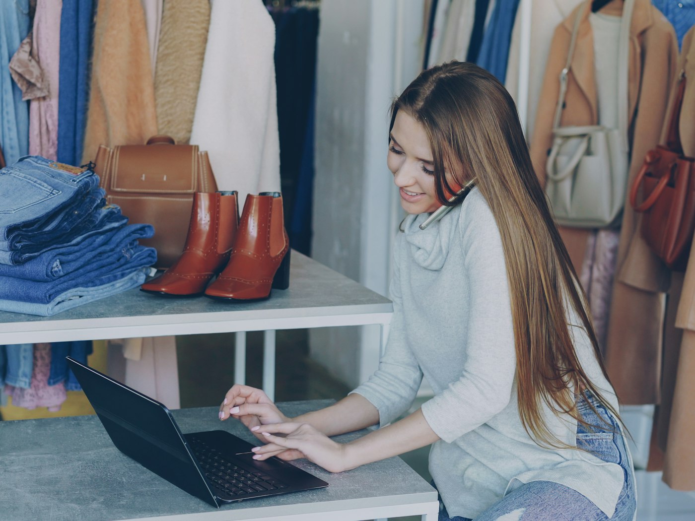A clothes shop owner on the phone, working on a laptop among her stock