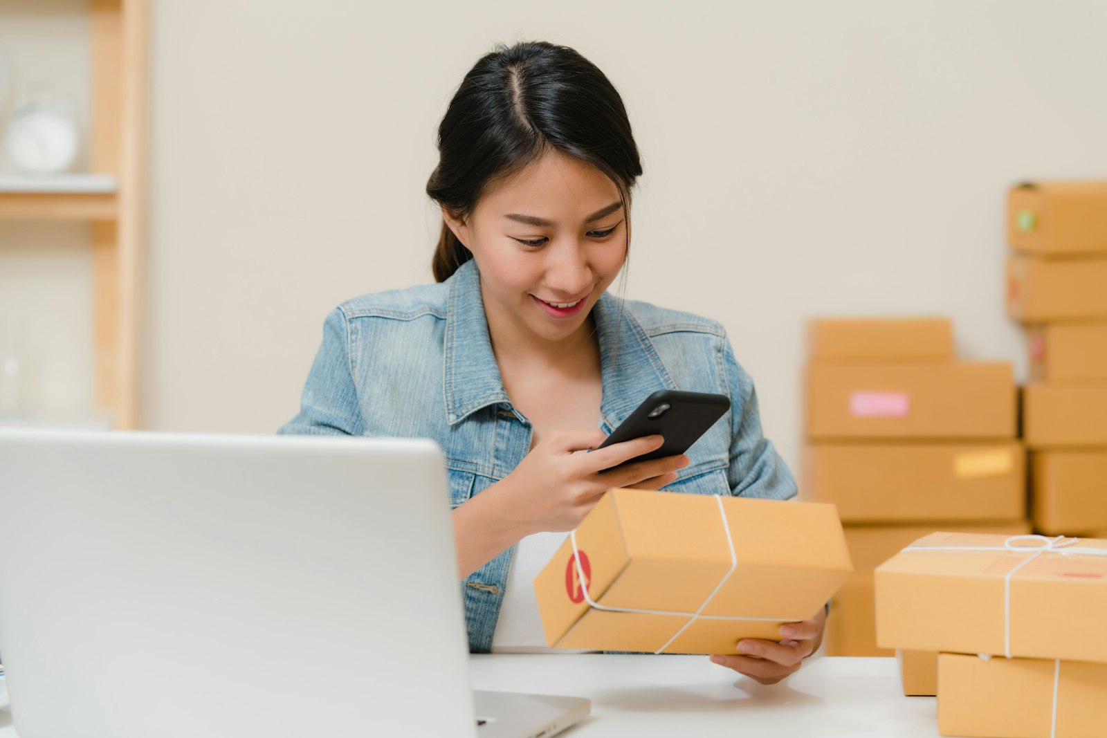 A seller checking a parcel on her phone beside a laptop and boxes